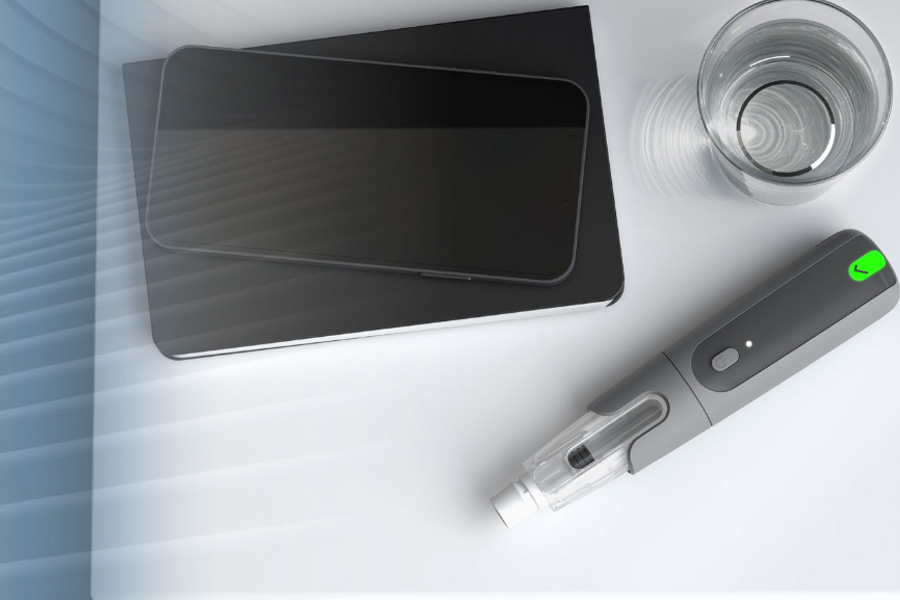 An image of a wooden tabletop, featuring a smartphone with a medical injection device app, the Aria Smart Autoinjector device and the packaging for this device