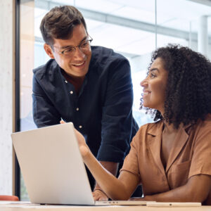 Two colleagues discuss automotive program management in an office. A female is sitting in front of a laptop while her male colleague stands next to her.