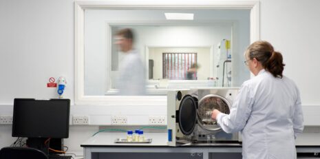 Phillips Medisize employee working in the pharmaceutical technology service lab, while we can see another employee walk past outside the window. Through the window, another corridor shows another employee in a neighbouring room.