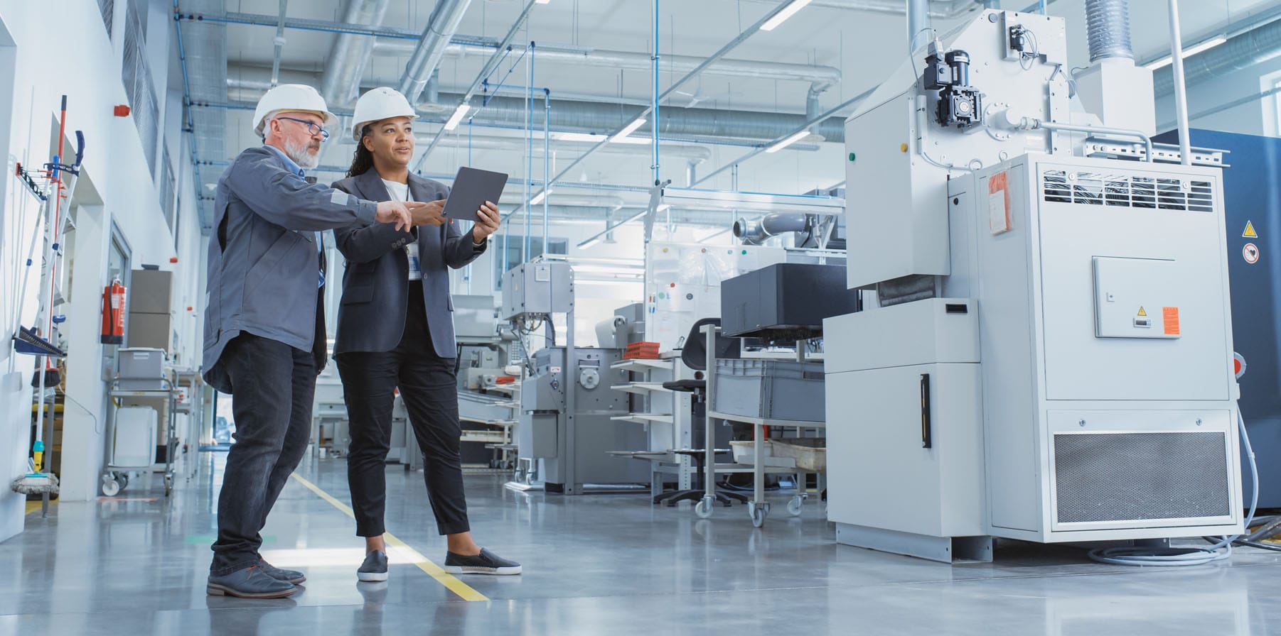 Portrait of two employees in hard hats aa manufacturing plant discussing lifecycle management using a tablet computer.